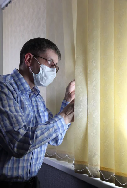 Man in a medical face mask looks out the window through the blinds — Stock Photo, Image