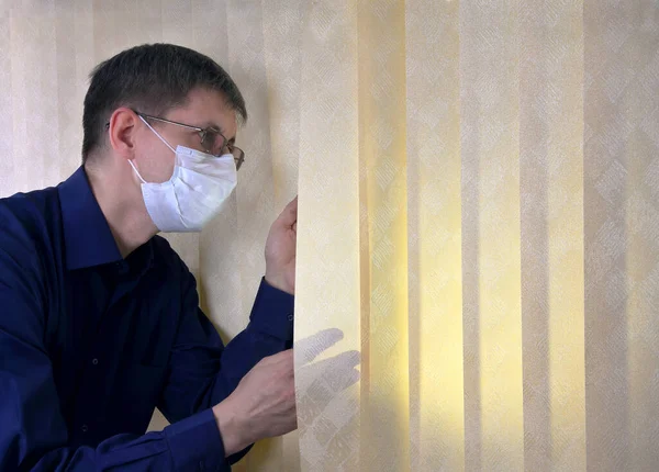 Man in a medical facial mask peeks out through the blinds — 스톡 사진