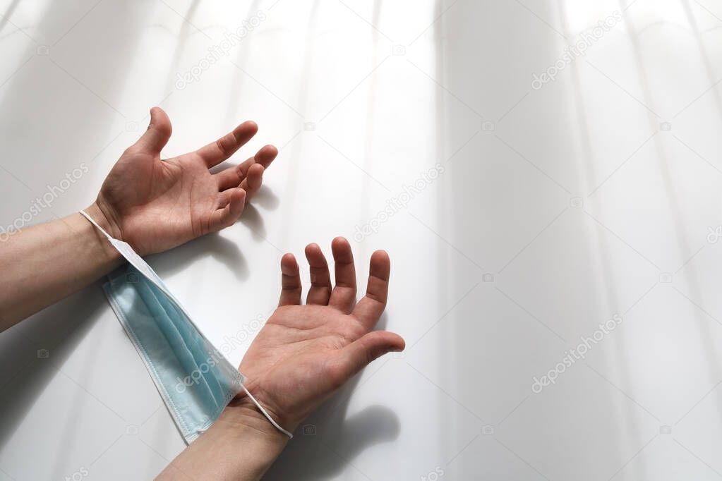 Hands shackled in a medical mask and lowered in despair. Against a light background in the form of a shadow from the prison bars with copy space