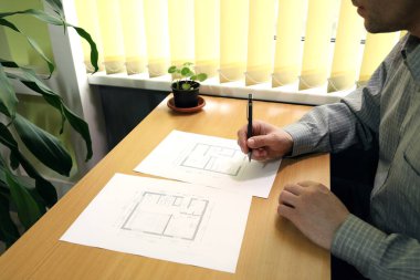 Bir adam pencerenin yanındaki masada oturur ve mimari kat planına notlar alır.