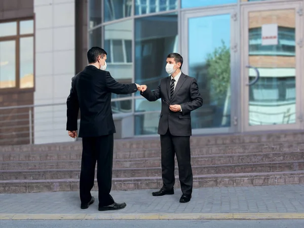 Two businessmen in medical face masks stand on the street near the steps at the entrance to the building. They practice greeting bump fists, abandoning the traditional handshake