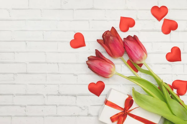 Fiori rossi tulipani, cuori e scatola regalo con fiocco rosso su uno sfondo bianco di muro di mattoni, primavera e vacanze, Festa della Donna . — Foto Stock