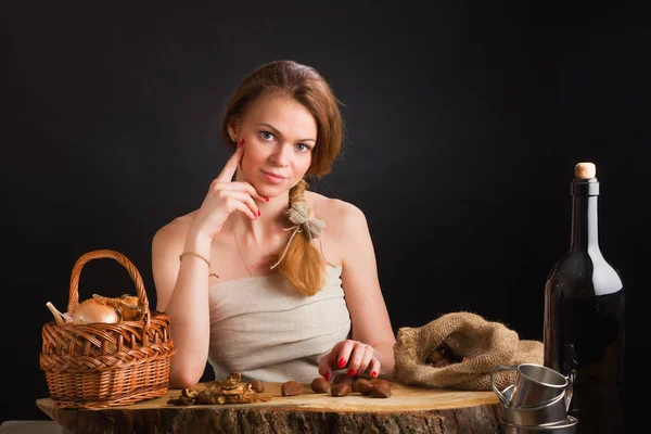 La muchacha joven hermosa en el vestido del lienzo se sienta a la mesa de roble sobre la cesta con la cebolla y el ajo, las setas secas las castañas Fotos De Stock Sin Royalties Gratis