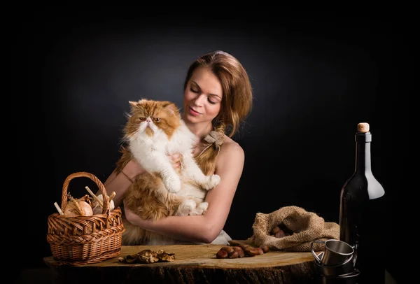 Het mooie meisje in een sundress van doek zit op een eiken tafel met opvallende rode Perzische kat op handen in de buurt van mand uien, gedroogde paddestoelen en kastanjes tas — Stockfoto