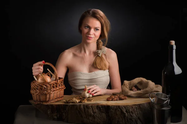 La joven hermosa en un vestido de tela se sienta en una mesa de roble con la cabeza de ajo en la mano cerca de la cebolla canasta y castañas bolsa Imagen De Stock