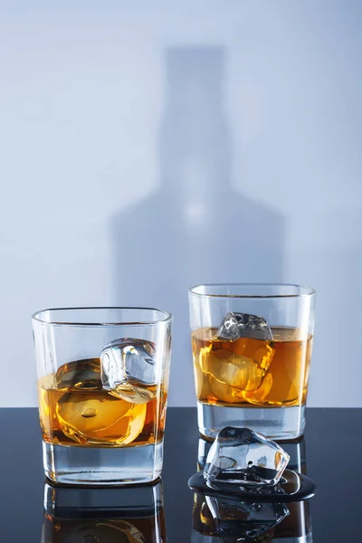 Dos vasos de whisky y hielo sobre un fondo claro con una sombra de una botella Fotos De Stock