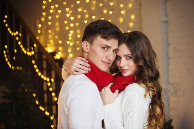 kucaklayan Noel ışıkları arka plan üzerinde poz genç mutlu çift beyaz kazak ve kırmızı eşarp