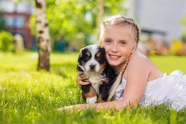 緑の草の上に横たわる黒のかわいい子犬を抱いて金髪少女 — ストック写真