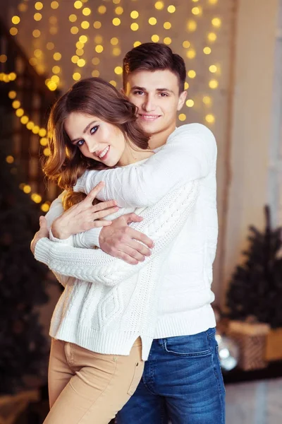 クリスマスの照明で部屋に立っている白いセーターでポーズを抱いて愛の若いカップル — ストック写真