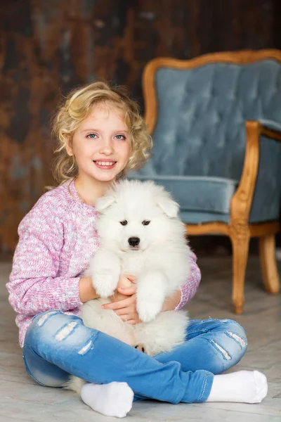 居心地の良い部屋で床に座って白いふわふわ犬と遊ぶ金髪少女 — ストック写真