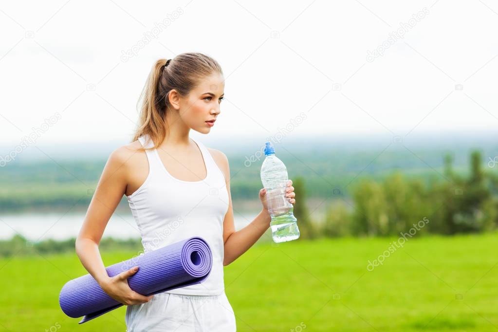 portrait of young sporty woman holding bottle of water and yoga mat after workout outdoor