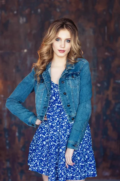 Young blonde woman with wavy hair wearing bright blue dress and denim jacket posing at camera