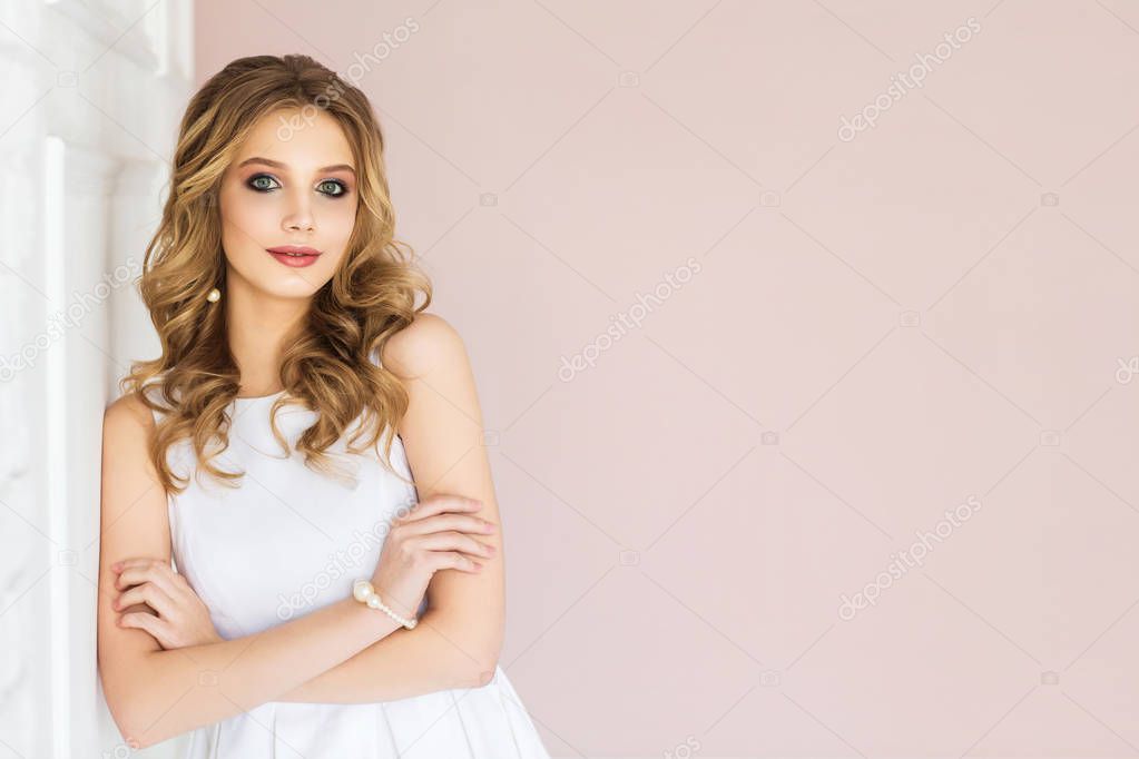 Portrait of young blonde woman with wavy hair wearing white dress posing at camera