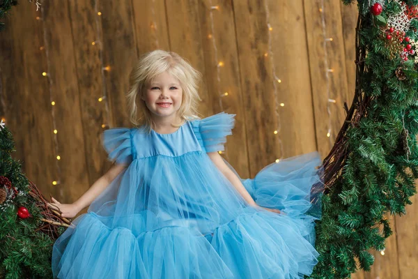 穿着蓝色连衣裙的迷人小女孩坐在圣诞花环上 — 图库照片
