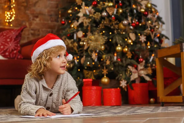 Little curly boy writes letter to Santa near christmas tree