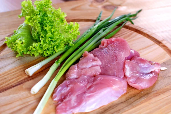 Slices of meat lie on a wooden board, with lettuce and chives. Sliced beef meat on a wooden table. Sliced meat with green onions and salad. Poultry meat pieces.