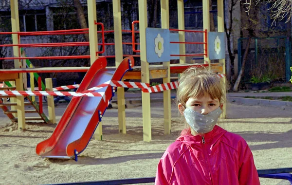 A little girl in a medical mask is not allowed to play in the playground. Little girl walks in a medical mask. A masked child is walking during quarantine. Indoor playground.