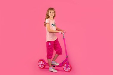 Pembe motosikletli küçük bir kız, pembe arka planda. Pembe tişörtlü ve şortlu güzel bir kız elinde bir scooter tutuyor. Motosikletli kasklı küçük bir kız. Scooter 'ı olan çocuk.