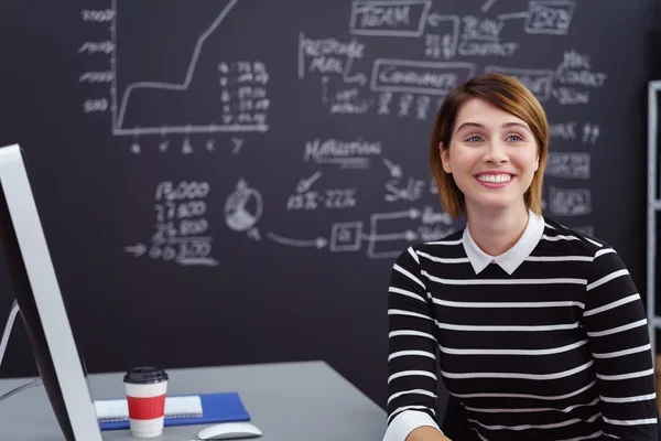 Woman sitting at desk with chalkboard behind her — Stockfoto