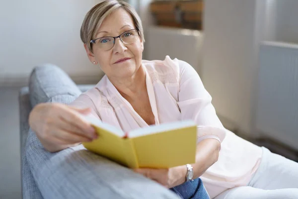 Femme âgée assise sur un canapé avec un livre — Photo