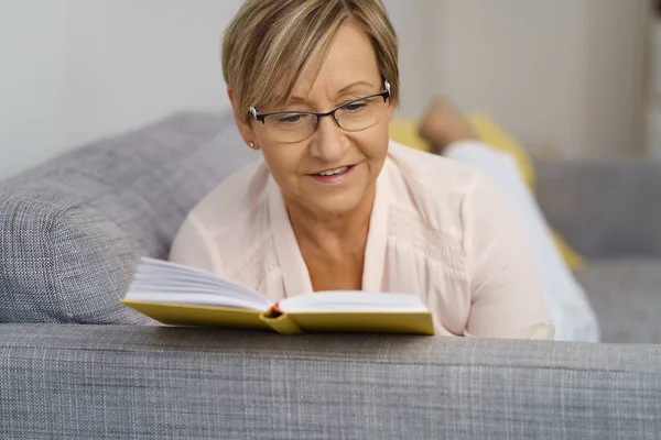 Sênior mulher leitura livro no sofá — Fotografia de Stock