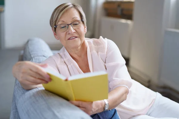 Sênior mulher leitura livro no sofá — Fotografia de Stock