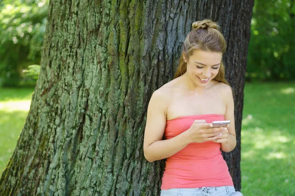 Mujer joven de pie junto al árbol con teléfono móvil — Foto de Stock