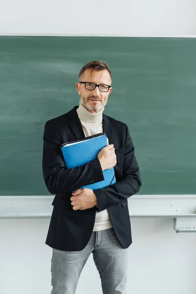 自信的聪明的男老师或教授站在教室的空白黑板前 手里拿着一个笔记夹在他的胸口 — 图库照片