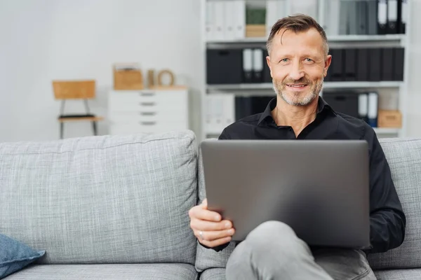 매력적인 남자가 소파에 미소를 카메라에 집에서 노트북으로 편안한 — 스톡 사진