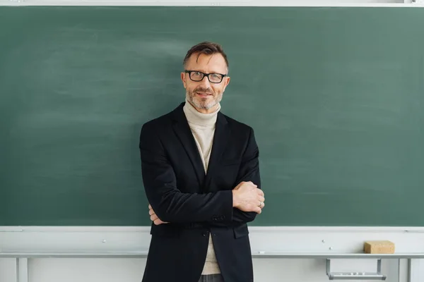 自信的中年男教师或教授站在一个空白的粉笔板前折叠手臂微笑 — 图库照片