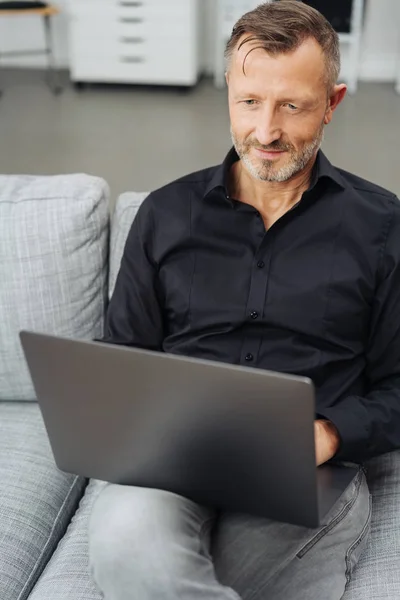 一个人在家里的沙发上使用笔记本电脑在他的膝上平衡它在屏幕上读数据在关闭视图 — 图库照片