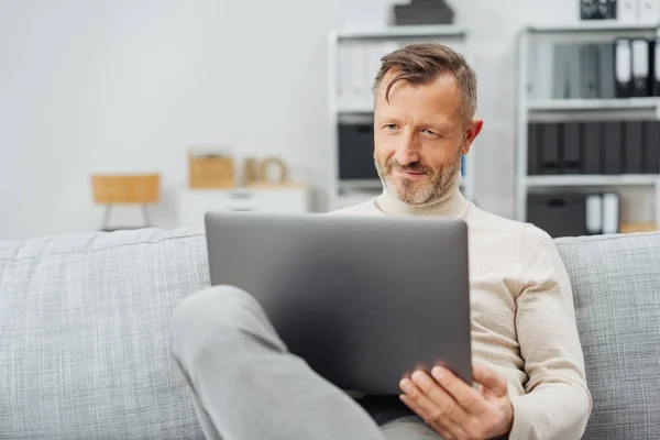 有吸引力的中年男子坐在舒适的沙发上 用笔记本电脑 高兴的微笑 他在家里放松 — 图库照片