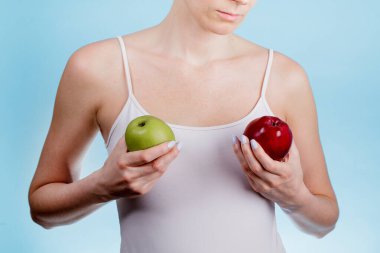 Ellerinde farklı renkte iki elmayla göğsünü kaplayan beyaz bir kadının yakın çekimi. Sağlıklı bir yemek fikri.