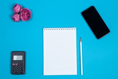 Tablet, akıllı telefon, hesap makinesi, çiçekler ve mavi arka planda beyaz boş not defteri olan ofis masası. Metin için üst görünüm ve kopyala