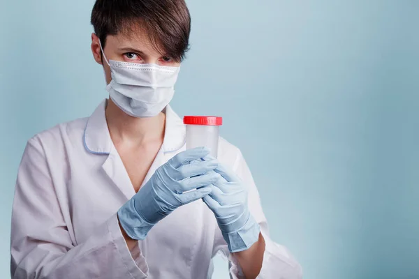 Tıbbi maskeli kadın doktor mavi arka planda boş bir test kavanozu tutuyor.