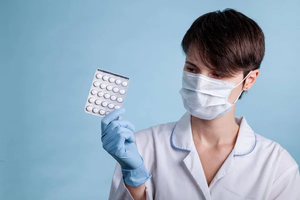 A young attractive female scientist in a mask holds a package of pills with gloved fingers on a blue background