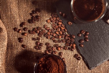 Çuval bezinin üstünde şeffaf cam bir fincanda sade kahve. Şekerin, kahve çekirdeğinin ve öğütülmüş kahvenin yanında. Vintage