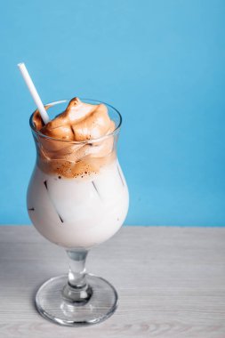 Dalgona Koreli buzlu kahve, beyaz ahşap bir masada buzlu kremalı, modaya uygun bir kahve. Mavi arka planda modaya uygun kahve içeceği