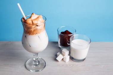 Dalgona Koreli buzlu kahve, beyaz ahşap bir masada buzlu kremalı, modaya uygun bir kahve. Mavi arka planda modaya uygun kahve içeceği