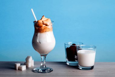 Dalgona Koreli buzlu kahve, beyaz ahşap bir masada buzlu kremalı, modaya uygun bir kahve. Mavi arka planda modaya uygun kahve içeceği