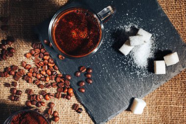 Çuval bezinin üstünde şeffaf cam bir fincanda sade kahve. Şekerin, kahve çekirdeğinin ve öğütülmüş kahvenin yanında. Yukarıdan görünüm