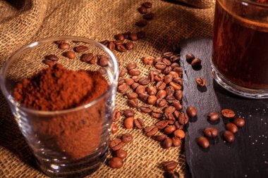 Taştan siyah bir stantta, cam bir fincanda koyu kahve eşliğinde çuval bezinin arka planında. Şeker, kahve çekirdeği ve kahvenin yanında..