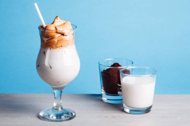Dalgona Koreli buzlu kahve, beyaz ahşap bir masada buzlu kremalı, modaya uygun bir kahve. Mavi arka planda modaya uygun kahve içeceği