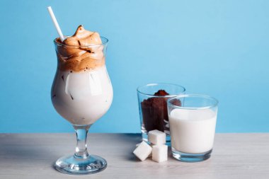 Dalgona Koreli buzlu kahve, beyaz ahşap bir masada buzlu kremalı, modaya uygun bir kahve. Mavi arka planda modaya uygun kahve içeceği