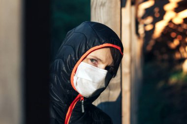 Yüzünde tıbbi maske olan üzgün bir genç kadının portresi ve sadece saklandığı yerden dışarı bakan bir başlık..