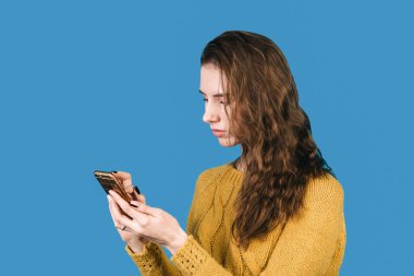 Siyah saçlı, gülümseyen genç bir kadının portresi mavi arka plandaki akıllı telefonuna bakıyor. Cep telefonunu kontrol eden bir kadın. İletişim konsepti.