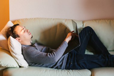 Laptopla çalışan ve evdeki kanepede yatan yakışıklı genç adam. Genç ve çekici adam evden çalışıyor. Acelesi ya da endişesi yok. Dizüstü bilgisayar ve modern teknolojiyle serbest çalışıyor..