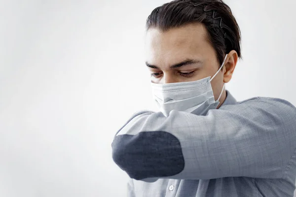 Sick young man in a medical face mask is coughing into his sleeve or elbow to prevent spread Covid-19, Corona virus. Coronavirus quarantine. Young man sneezing into his elbow on white backround.