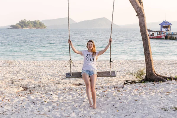 A girl swinging on a swing and looks at the camera on a tropical seashore of beach, Cambodia, Koh Rong. The swing has simple wood construction. Freedom, rest, vacation, travel concept. Copy space.