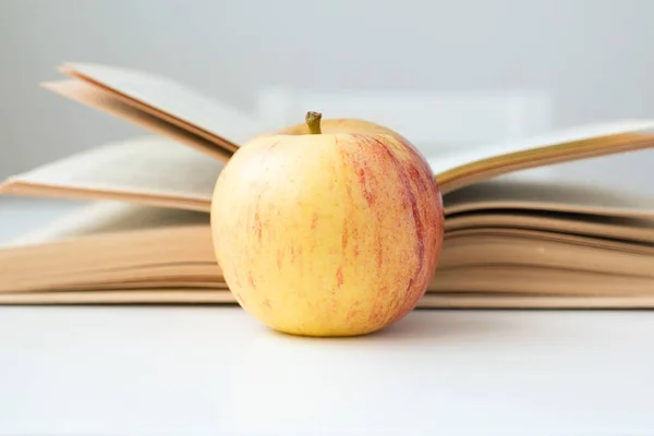 On the table is a book with an apple. A red-yellow apple is in front of a book. On the desk is an apple.